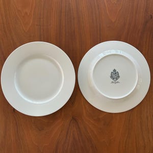 May include: Two white ceramic plates on a wooden surface. One plate is face up, and the other is face down, revealing a crest design on the underside. The plates have a simple, elegant design.