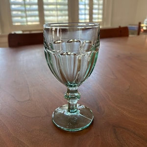 May include: A clear, vintage-style water goblet with a decorative, ribbed design on the bowl. The stem features a small, round detail. The glass sits on a round base. The goblet is made of clear glass with a slight green tint.