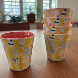 May include: A set of six yellow cups with a botanical pattern of potted plants in various shapes and sizes. The cups have a ribbed texture and a coral-colored interior. The cups are stacked and arranged on a wooden table.