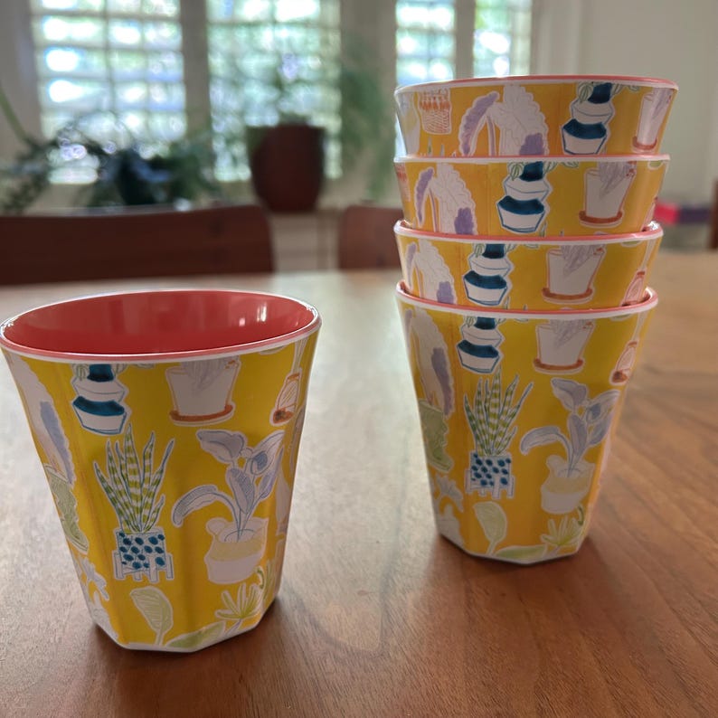 May include: A set of six yellow cups with a botanical pattern of potted plants in various shapes and sizes. The cups have a ribbed texture and a coral-colored interior. The cups are stacked and arranged on a wooden table.