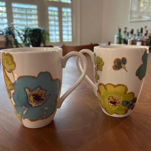 May include: Two white ceramic mugs featuring a colorful floral pattern. The left mug displays a large blue flower with yellow and brown accents, while the right mug features a prominent lime green flower with blue and brown details and a curved handle.