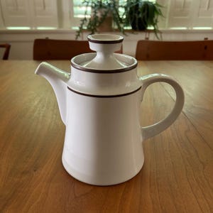 May include: A white ceramic teapot featuring a minimalist cylindrical body, a curved handle, and a short spout. The lid is topped with a small, knob-like handle, and the entire piece is accented with thin, dark brown horizontal trim lines.