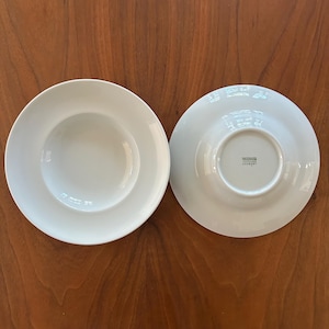 May include: Two white ceramic soup bowls are displayed on a wooden surface. One bowl is right-side up, showing the interior, while the other is upside down, revealing the brand name. The bowls have a simple, elegant design.