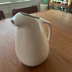 May include: A beige ceramic pitcher with a curved handle and a spout. The pitcher has a smooth, matte finish and a rounded base. The pitcher is sitting on a wooden table.