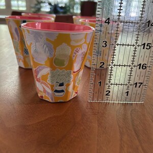 May include: Set of six yellow cups with a botanical pattern of plants and flowers. Each cup has a pink interior and is approximately 3 inches tall. The cups are arranged on a wooden surface.