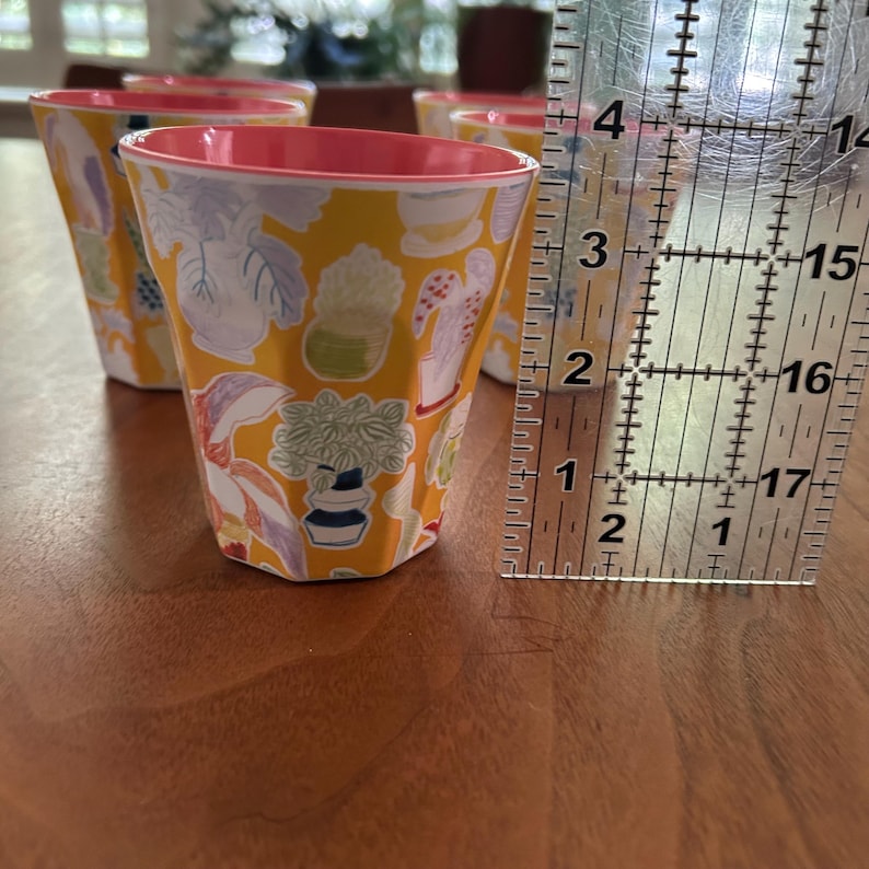May include: Set of six yellow cups with a botanical pattern of plants and flowers. Each cup has a pink interior and is approximately 3 inches tall. The cups are arranged on a wooden surface.