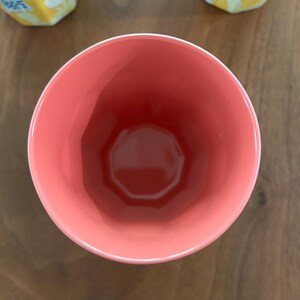 May include: A coral-colored ceramic cup with a geometric design. The cup has a wide opening and a faceted exterior. The interior is a darker shade of coral. The cup is sitting on a wooden surface.