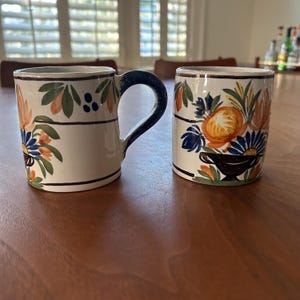 May include: Two ceramic mugs with floral designs. One mug has a handle with a dark blue color, while the other is handle-less. Both mugs feature a cream-colored base with orange, blue, and green floral patterns and a dark brown rim.