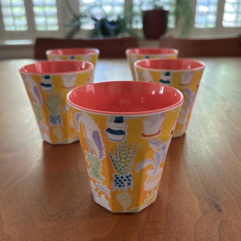 May include: A set of five yellow cups with a coral-colored interior. The cups feature a botanical design with various potted plants and flowers. The cups are arranged on a wooden table, showcasing their unique design.
