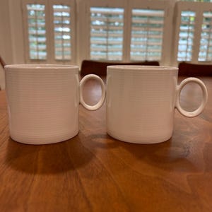 May include: Two white ceramic mugs with horizontal ribbed detailing. Each mug has a rounded handle and a cylindrical shape. The mugs are placed on a wooden surface, with natural light coming from the background.
