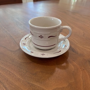 May include: A white teacup and saucer set with a burgundy floral design. The cup has a handle and sits on a matching saucer. The set is made of ceramic and is suitable for serving tea or coffee.