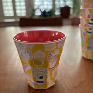 May include: A yellow cup with a red interior and a white rim. The cup is decorated with a pattern of various potted plants in white, green, and blue. A stack of similar cups is visible in the background.