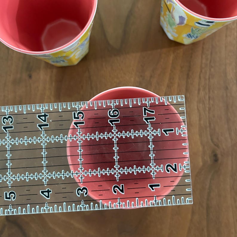 May include: A close-up of a red cup with a pink interior, measuring approximately 3 inches in diameter. The cup is placed on a wooden surface, with a clear ruler positioned across its top for measurement. Two other cups are visible.