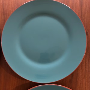 May include: Two teal-colored ceramic plates with a thin orange rim. The plates have a simple, modern design and are suitable for everyday use. The plates are round and have a slightly raised edge.