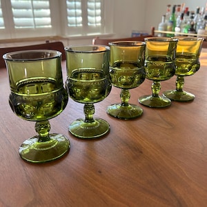 May include: Five vintage green-tinted glass goblets with a decorative pattern around the bowl and on the base. The goblets have a short stem and a wide, round base. The glasses are arranged on a wooden table.