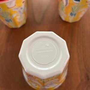 May include: Yellow and white ceramic mugs with a floral design. The mugs have a unique, faceted shape and are branded with "by ANTHROPOLOGIE" on the bottom. The mugs are decorated with colorful illustrations of plants and flowers.