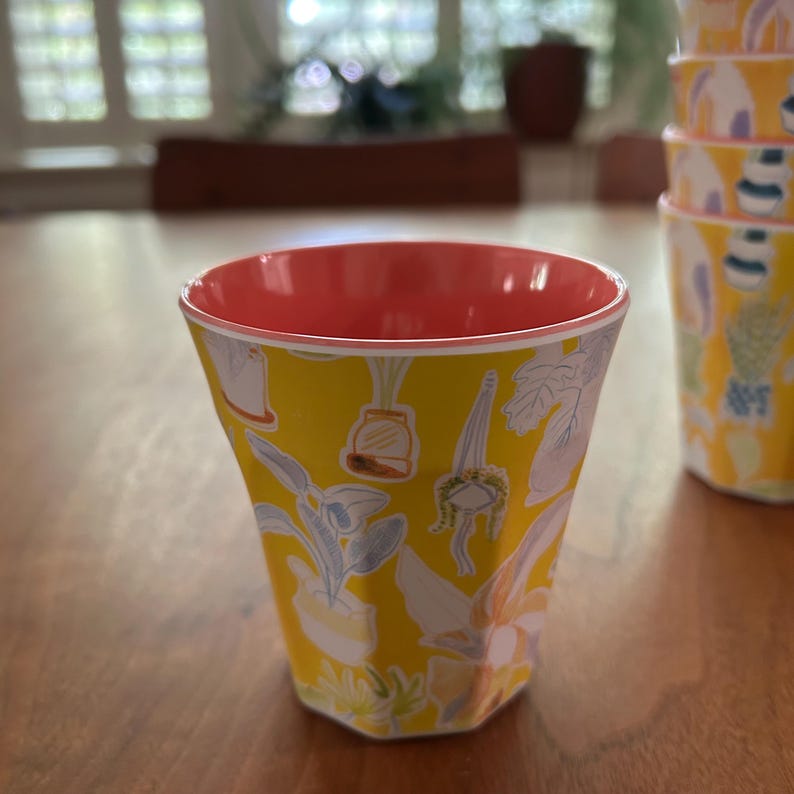 May include: A yellow cup with a coral interior and a botanical pattern featuring various potted plants. The cup has a unique, faceted shape and is part of a set. The design includes white, gray, and green plant illustrations.