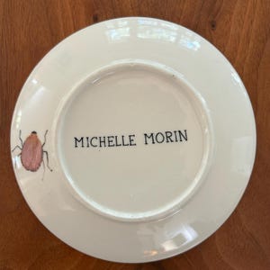以下が含まれることがあります： ピンクと茶色の昆虫のデザインが描かれた白いセラミックプレート。 プレートには「MICHELLE MORIN」の文字が黒で印刷されています。
