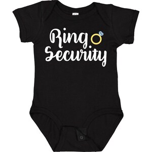 May include: Black baby bodysuit with the words "Ring Security" in white script. A yellow ring with a blue diamond is to the right of the text. The bodysuit has short sleeves and a snap closure.