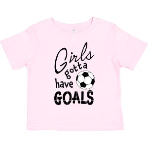 May include: A light pink short-sleeved t-shirt featuring black text that reads Girls gotta have GOALS, with a black and white soccer ball graphic replacing the letter O in the word goals. The design is centered on the front of the shirt.