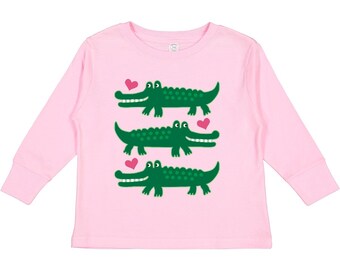 May include: A light pink long-sleeved shirt featuring three green cartoon alligators, each with a pink heart. The alligators are smiling. The shirt is designed for toddlers.