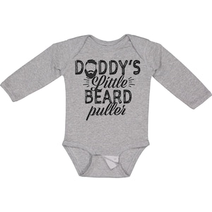 May include: A gray long-sleeved baby bodysuit with the text "DODDY'S Little BEARD puller" in black. The design includes a beard graphic above the words. The bodysuit has a snap closure at the bottom.