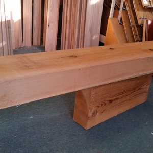 May include: A large, unfinished, light brown wooden beam, approximately 8 feet long, rests on a smaller, unfinished, light brown wooden block. The beam is smooth and has a natural wood grain.
