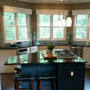May include: A kitchen island with a dark blue base and a black granite countertop featuring a built-in gas cooktop. Two black leather stools with wooden legs are positioned at the island. Three white pendant lights hang above the counter.