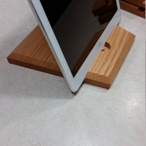 May include: A wooden tablet stand made of light brown oak. The stand is designed to hold a tablet upright, with a cutout for the tablet's camera.