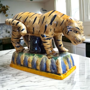 May include: A ceramic tiger figurine with a tan body and black stripes. The tiger is standing on a base with blue, yellow, and green accents. The figurine is a decorative item.