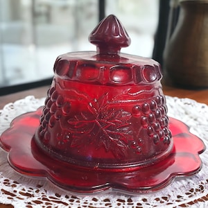 May include: A red glass butter dish with a grapevine design. The dish has a domed lid with a knob on top. The dish is sitting on a white lace doily.