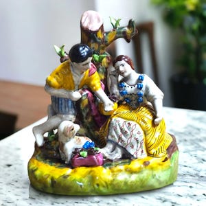 May include: A porcelain figurine depicting a couple and a small dog. The man is wearing a yellow shirt and blue pants, the woman is wearing a yellow dress with blue and red accents, and the dog is white with brown spots. The figurine is set against a green and brown background.