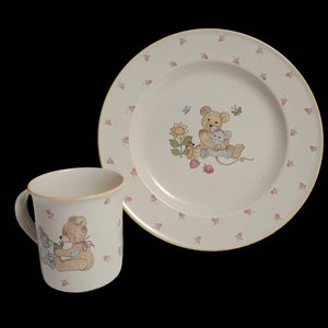 May include: A set of children's dishes featuring a brown teddy bear and a gray mouse. The plate and mug are white with a pink and yellow floral pattern.