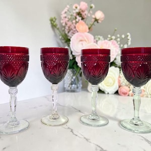 May include: Four ruby red wine glasses with intricate cut glass patterns. The glasses have clear stems and bases, and are arranged on a white surface. A bouquet of pink and white flowers is in the background.
