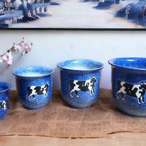 May include: Four blue ceramic flower pots with a white cow design. The pots are different sizes and are arranged in a row from largest to smallest.