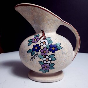 May include: A white ceramic pitcher with a floral design in shades of blue, purple, green, and white. The pitcher has a brown rim and a signature that reads "A.Puyois".