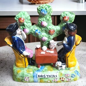 May include: A ceramic figurine depicting two people sitting in chairs, playing cards under a tree with pink flowers. The figurine is painted in bright colors and has the text "DRS YNTAX" on the base.
