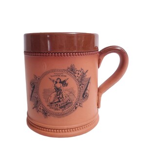 May include: A vintage-style, brown ceramic mug with a handle. The mug features a detailed black and white illustration with the text "Harrods" and "Omnibus Unique World". The rim and handle are a darker shade of brown.