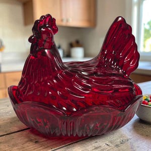 May include: A ruby red glass hen-shaped dish with a detailed comb and tail feathers. The dish is sitting on a wooden surface. The hen is a decorative kitchen item.