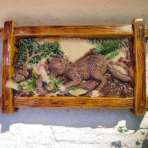 May include: A rectangular ceramic wall plaque with a relief scene of a brown animal with a long tail, possibly a fox or a weasel, attacking a bird. The scene is framed by a wooden-look border.