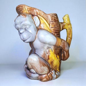 May include: A vintage ceramic pitcher featuring a monkey figure. The monkey is depicted in shades of brown, tan, and white, with a basket-like structure on its back. The handle is designed to resemble a bamboo stalk.