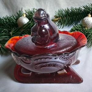 May include: A red glass candy dish shaped like a sleigh with a red glass bird figurine on top. The dish has a detailed design with swirls and a scalloped edge.