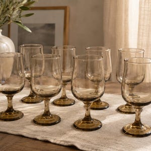 May include: A set of eight amber-colored wine glasses with a classic stem design. The glasses are arranged on a neutral-toned linen tablecloth. The glasses have a rounded bowl and a sturdy base.