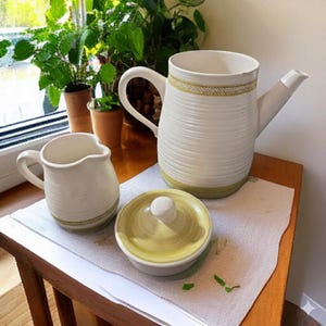 May include: A white ceramic pitcher, creamer, and lidded bowl set on a wooden table. The pitcher has a long spout and a handle. Each piece has a green and yellow geometric design around the rim and base. The creamer and bowl are smaller.