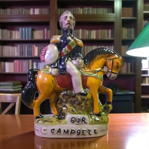 May include: A ceramic figurine of a man in a military uniform riding a horse. The figurine is painted with yellow, black, red, and blue colors. The base of the figurine reads "SIR CAMPBELL". The background shows a library with bookshelves.