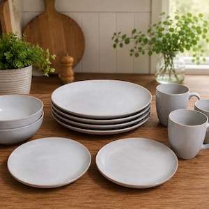 May include: A collection of light gray ceramic tableware, including plates, bowls, and mugs, arranged on a wooden surface. The plates are stacked, and the bowls are nested. The mugs have handles. The set is designed for dining.