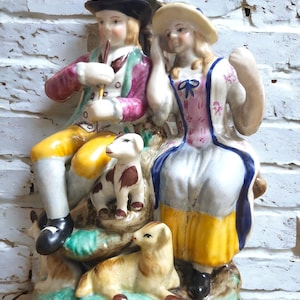 Puede incluir: Una figurita de porcelana vintage de una pareja con ropa tradicional, sosteniendo una pipa y un abanico, con dos pequeños perros blancos y marrones a sus pies. La figurita está colocada sobre una base verde y marrón.
