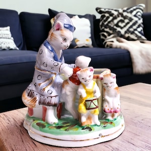 May include: A porcelain figurine of a cat family playing music. The father cat is wearing a blue and white nightgown and playing a violin. The mother cat is wearing a red dress and playing a trumpet. The baby cat is wearing a yellow dress and playing a drum.