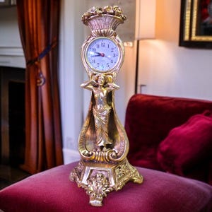 May include: A gold-colored decorative clock with a Roman numeral clock face. The clock features a sculpted female figure and floral accents. The clock is placed on a red velvet surface.