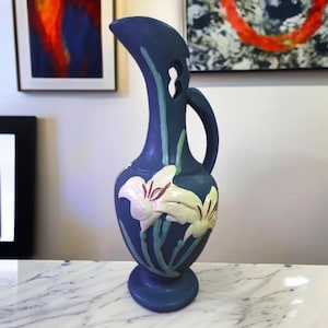May include: A navy blue ceramic vase with a handle and a unique curved top. The vase features white and yellow lily flower designs with red accents and green stems. The vase is approximately 12 inches tall.